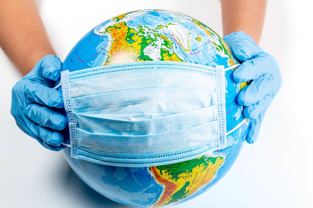 Image of two hands in medical gloves, holding a blue facemask over a globe of Earth.